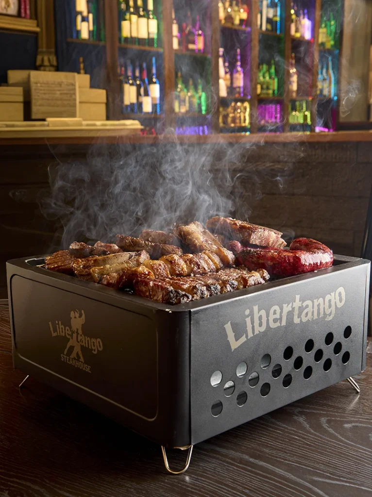 Parrillada Argenita - Sizzling assorted meats and sausages smoking on a Libertango Steakhouse tabletop grill in a dimly lit bar setting.